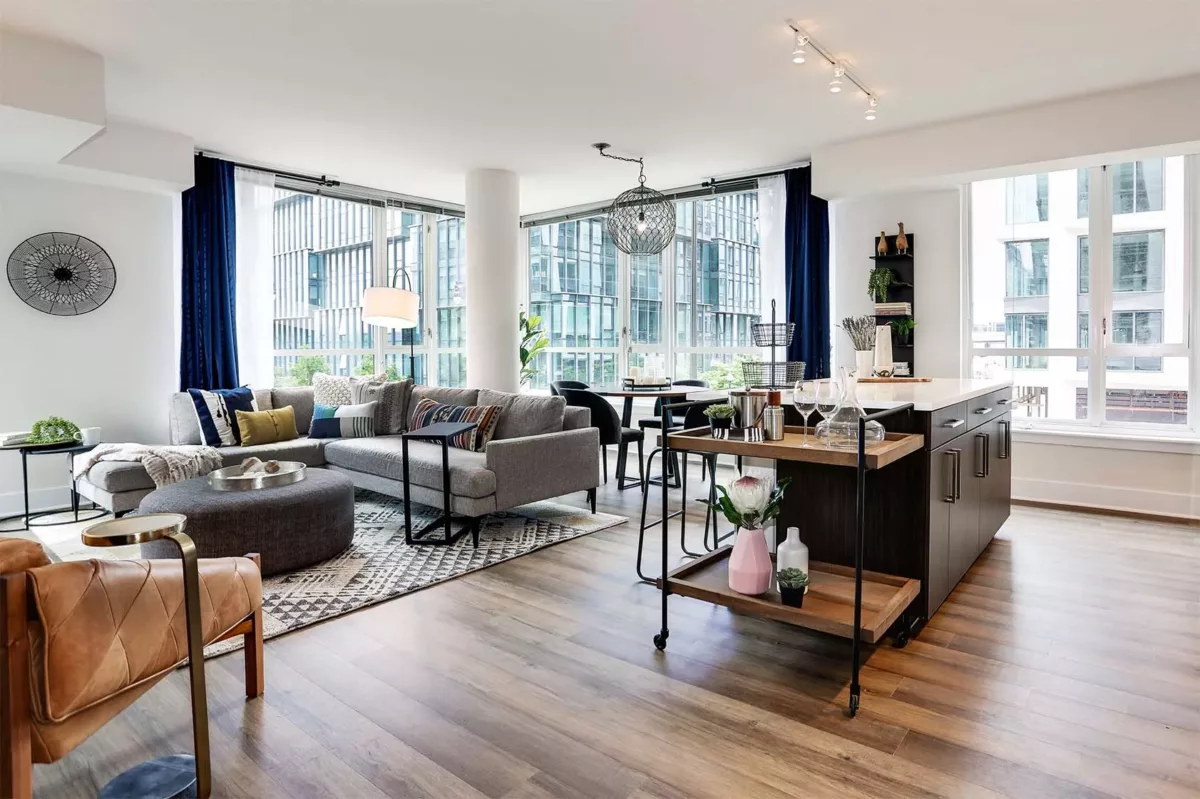 Open living room and kitchen with large windows, sectional sofa, island seating, and wood floors.