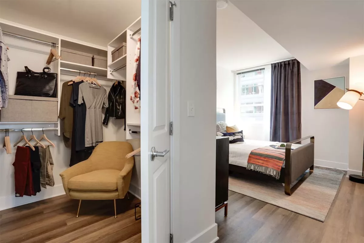 Walk‑in closet with open shelving, hanging clothes, accent chair, and adjacent bedroom.