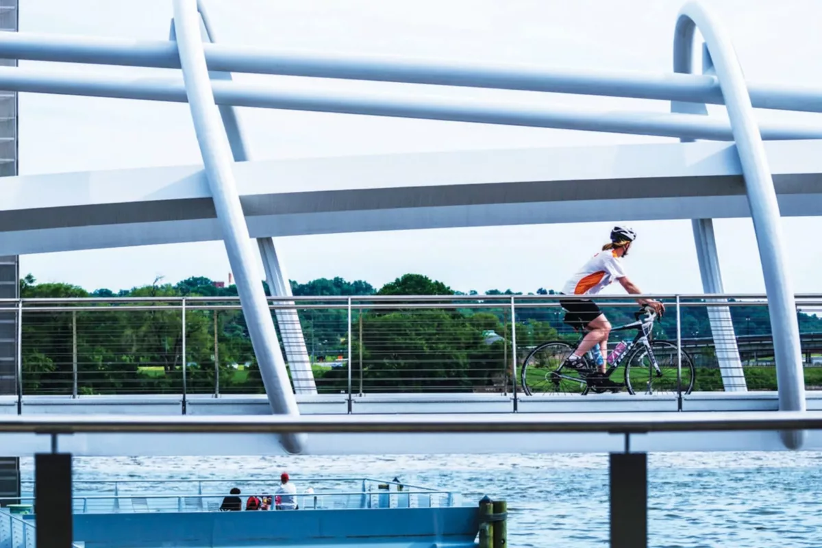 Pedestrian bridge with cyclist crossing over water and green surroundings.
