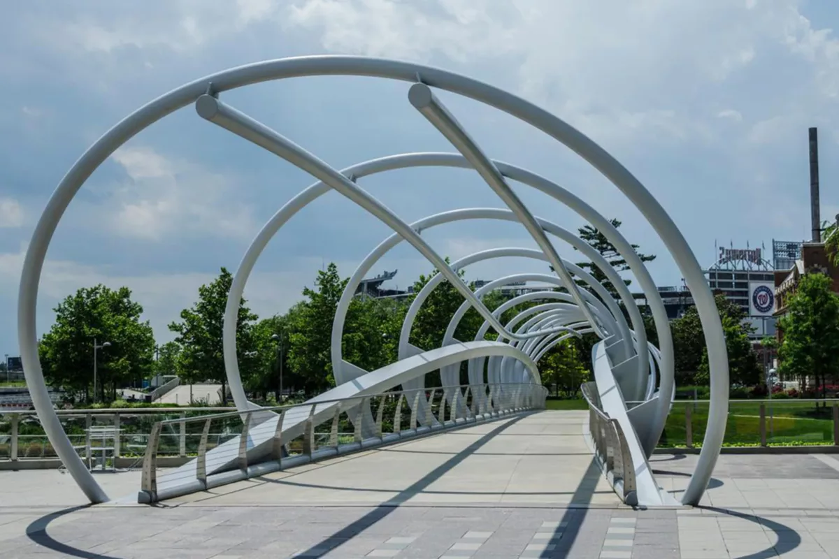 Curved pedestrian bridge with metal arches and landscaped surroundings.