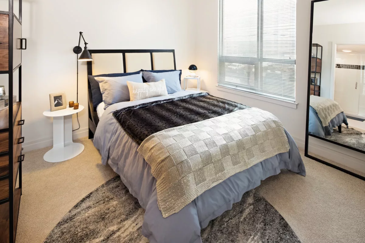 Bedroom with queen bed, side tables, floor mirror, neutral bedding, and window blinds.