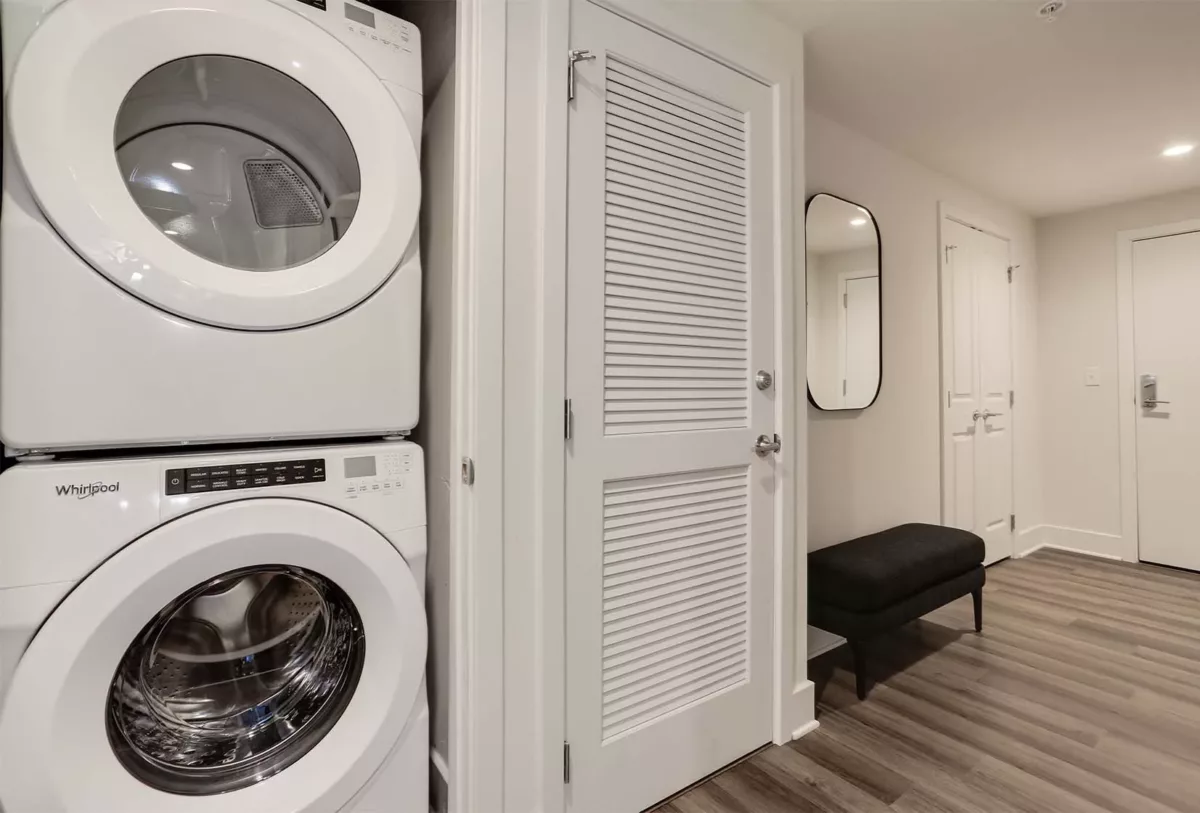 Stacked washer and dryer in hallway closet with bench seating and entry doors.